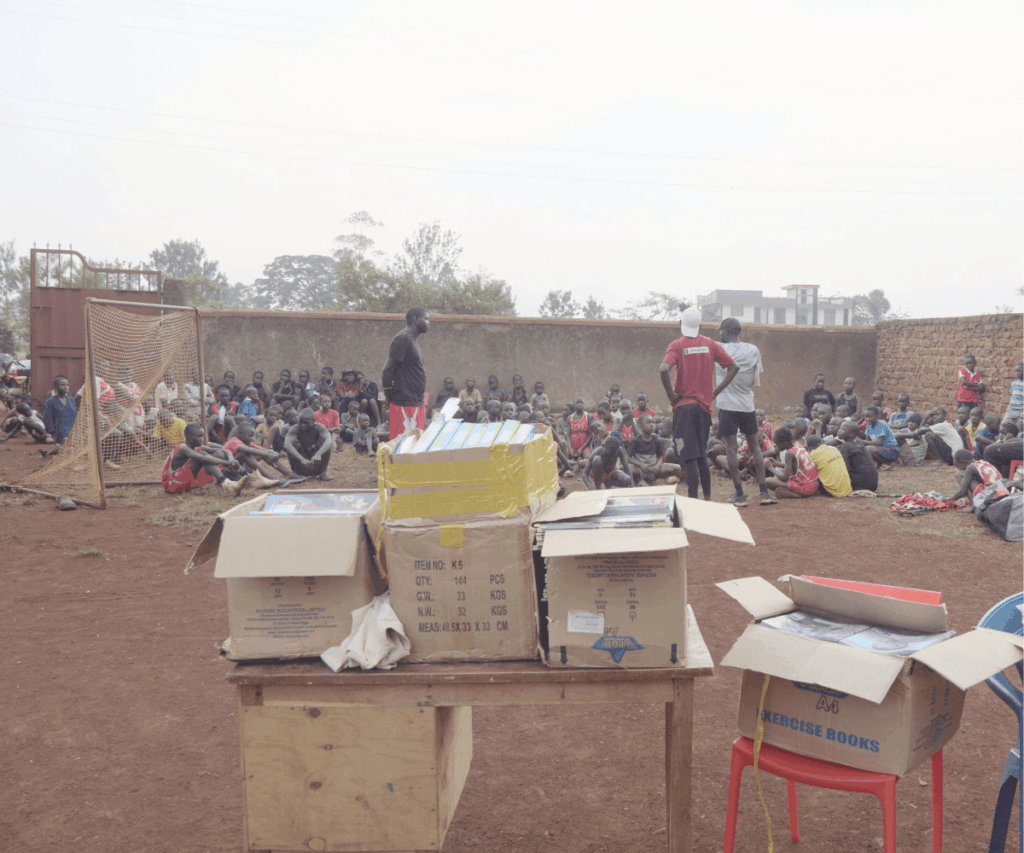 Large group of kids sitting next to a lacrosse goal as volunteers speak with them. Boxes of workbooks and supplies sit in front of them.